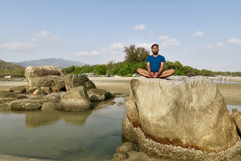 印度男子在岩石上练习蝴蝶/鞋匠姿势(Baddha Konasana)瑜伽姿势，水边，Palolem海滩，果阿，印度